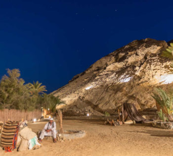 A man sitting beside his camel in the desert of El Gouna Egypt in the night while the mountains around the place