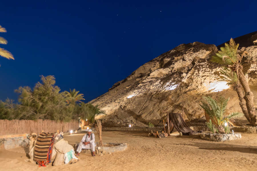 A man sitting beside his camel in the desert of El Gouna Egypt in the night while the mountains around the place