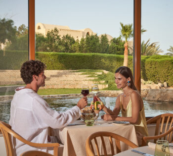 Couple sitting on wooden furniture in sunny outdoor area, cheering with wine glasses.