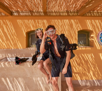 A man and a woman in diving suits and snorkeling masks laugh in a shaded outdoor setting.